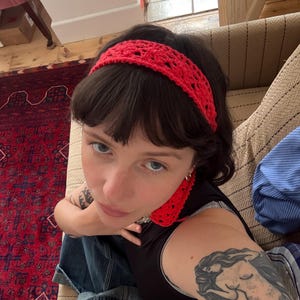 May include: A red crocheted headband with an openwork pattern. The headband is worn on dark hair. The person is wearing a black top and blue jeans. A red crocheted earring is visible.
