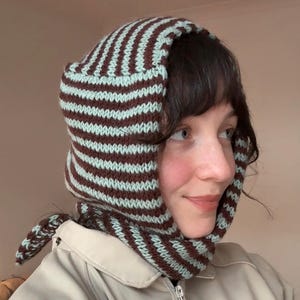 May include: A knitted hood in brown and light blue stripes. The hood is designed to cover the head and neck, with a tie closure. The knitted hood is worn over a beige jacket.