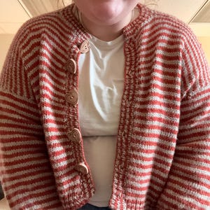 May include: A red and white striped knit cardigan with heart-shaped wooden buttons. The open-front sweater has a classic striped pattern and a cozy, handmade look. The cardigan is worn over a white t-shirt and blue jeans.