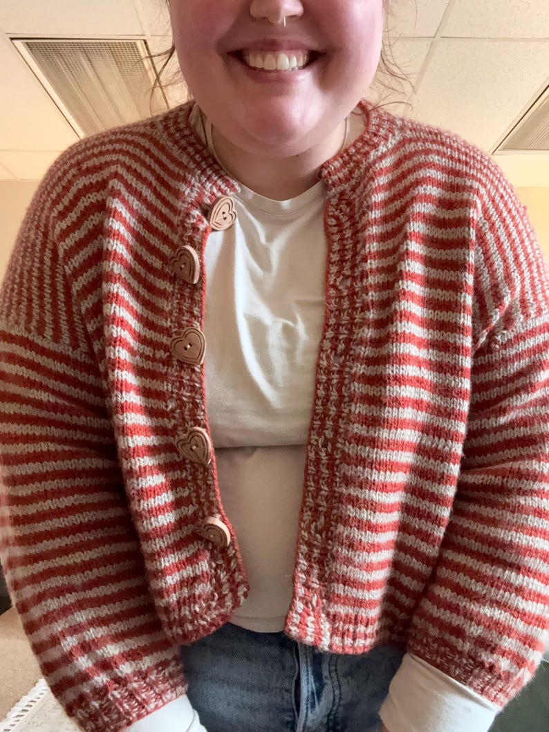 May include: A red and white striped knit cardigan with heart-shaped wooden buttons. The open-front sweater has a classic striped pattern and a cozy, handmade look. The cardigan is worn over a white t-shirt and blue jeans.