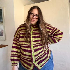 May include: A striped cardigan in shades of green and burgundy. The button-up sweater has a high collar and is worn with blue jeans. The cardigan features horizontal stripes and green buttons.