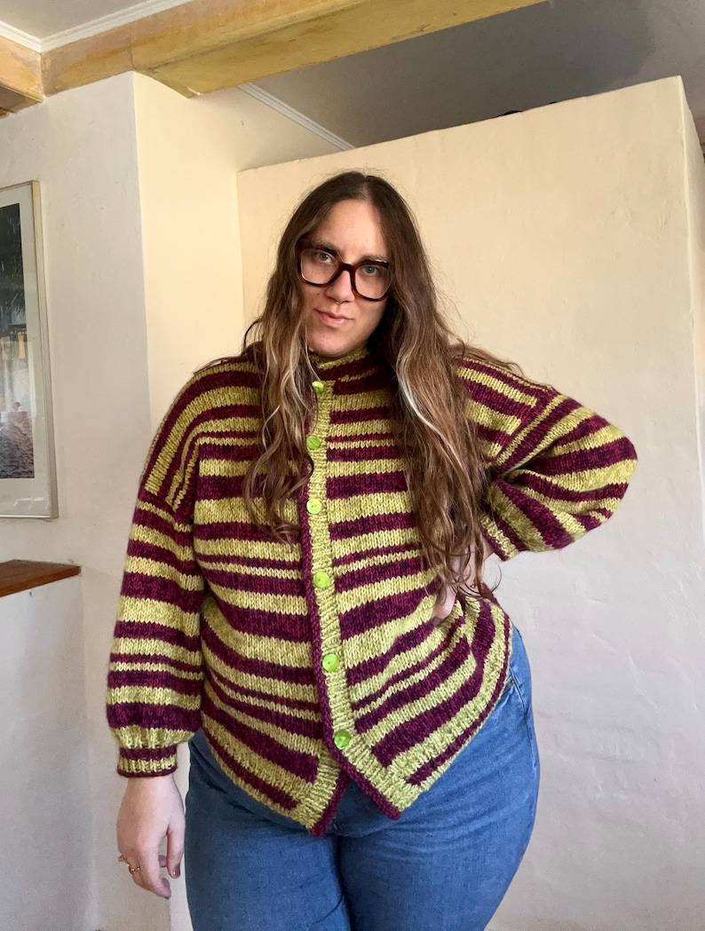 May include: A striped cardigan in shades of green and burgundy. The button-up sweater has a high collar and is worn with blue jeans. The cardigan features horizontal stripes and green buttons.