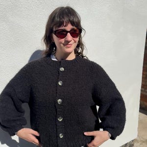 May include: A black, textured cardigan sweater with large, light-colored buttons. The sweater has a round neckline and long sleeves. The person in the image is wearing sunglasses and a white shirt underneath the sweater. The sweater is a casual, everyday garment.