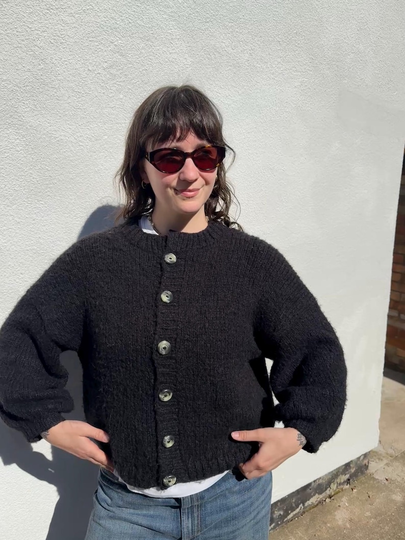 May include: A black, textured cardigan sweater with large, light-colored buttons. The sweater has a round neckline and long sleeves. The person in the image is wearing sunglasses and a white shirt underneath the sweater. The sweater is a casual, everyday garment.