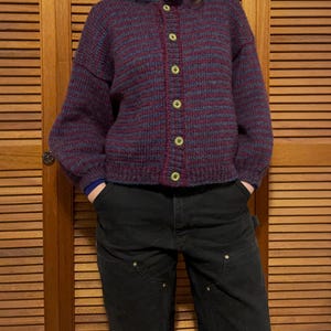 May include: A burgundy and blue striped knit cardigan with a button-up front. The sweater has long sleeves and a round neckline. The buttons are a light yellow color. The person is wearing black pants.