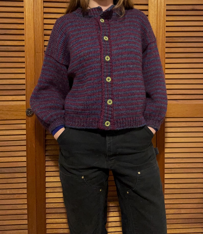 May include: A burgundy and blue striped knit cardigan with a button-up front. The sweater has long sleeves and a round neckline. The buttons are a light yellow color. The person is wearing black pants.
