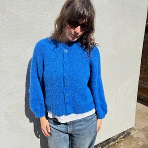May include: A vibrant blue cardigan with long sleeves and a button-up front. The cardigan is paired with light-wash denim jeans. The outfit is worn by a person wearing sunglasses.