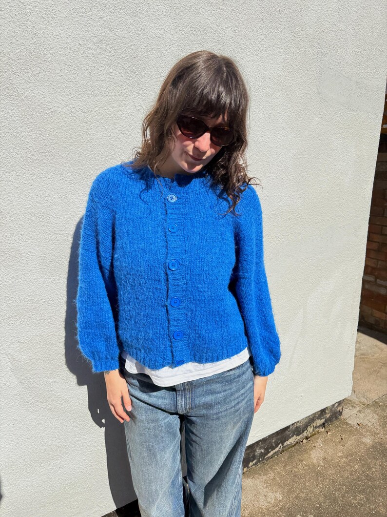 May include: A vibrant blue cardigan with long sleeves and a button-up front. The cardigan is paired with light-wash denim jeans. The outfit is worn by a person wearing sunglasses.