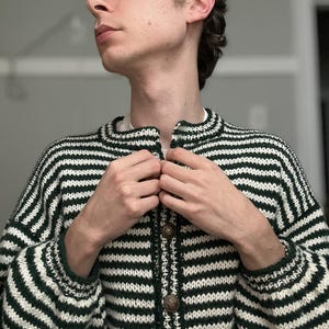 May include: A green and white striped cardigan with a button-up front. The sweater has a round collar and long sleeves. The buttons are a bronze color. The person in the image is wearing a green cap.