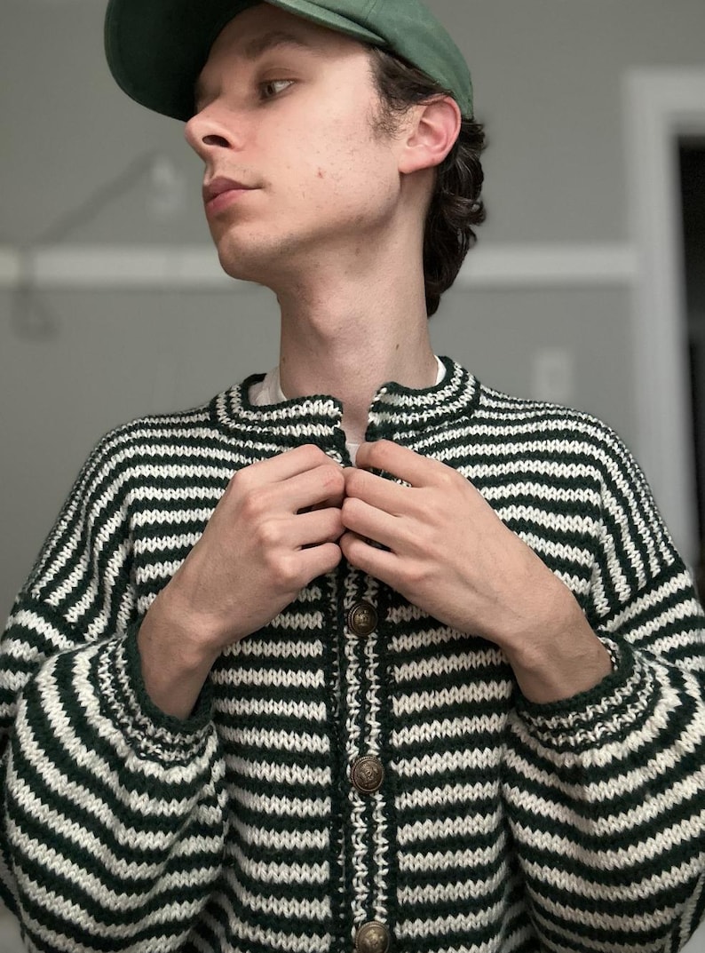 May include: A green and white striped cardigan with a button-up front. The sweater has a round collar and long sleeves. The buttons are a bronze color. The person in the image is wearing a green cap.