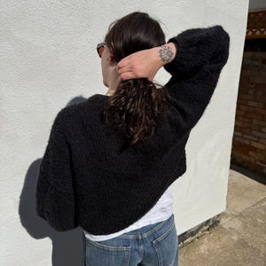 May include: A black, cropped knit sweater with long, voluminous sleeves. The sweater is worn over a white t-shirt and paired with blue jeans. The person is facing away from the camera, with their hair pulled back.