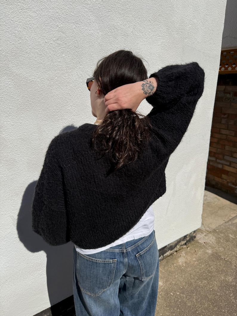May include: A black, cropped knit sweater with long, voluminous sleeves. The sweater is worn over a white t-shirt and paired with blue jeans. The person is facing away from the camera, with their hair pulled back.