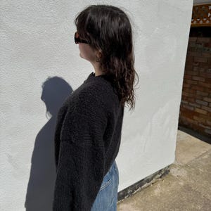 May include: A black knit sweater with long sleeves and a crew neck. The sweater is paired with blue jeans. The person is wearing sunglasses. The background is a white wall with a shadow.