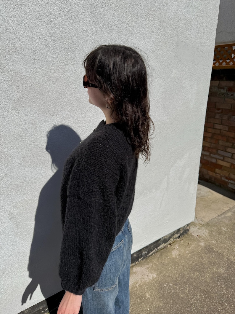 May include: A black knit sweater with long sleeves and a crew neck. The sweater is paired with blue jeans. The person is wearing sunglasses. The background is a white wall with a shadow.