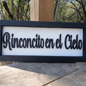 May include: A rectangular wooden sign with a black frame and a white interior. The sign features the phrase "Rinconcito en el Cielo" in black, cursive lettering. The sign is displayed outdoors.