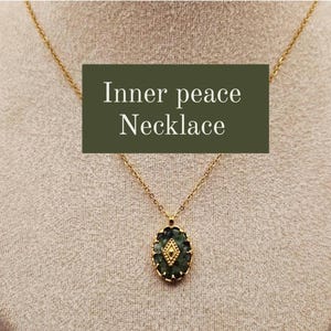 Op de afbeelding: Een goudkleurige ketting met een hanger met een donkergroene ovale steen met een goudkleurig ruitvormig accent. De ketting wordt getoond tegen een neutrale achtergrond. De tekst "Inner peace Necklace" staat in een donkergroene doos.
