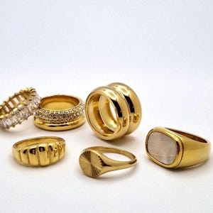 Könnte beinhalten: Ein Set aus sechs goldfarbenen Ringen. Die Ringe sind alle in verschiedenen Stilen, darunter ein Ring mit einem weißen Stein, ein Ring mit einer strukturierten Oberfläche und ein Ring mit einer Reihe kleiner Steine.