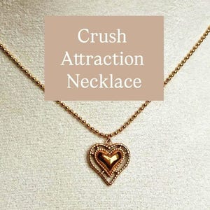 Puede incluir: Un collar dorado con un colgante en forma de corazón. El colgante tiene un diseño en capas con un corazón más pequeño en el interior. La cadena del collar está hecha de pequeñas cuentas redondas. El texto "Crush Attraction Necklace" está en la imagen.