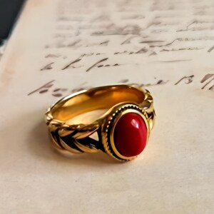 Könnte beinhalten: Ein goldfarbener Ring mit einem großen, ovalen, roten Edelstein. Der Ring hat ein detailliertes Design um das Band und die Edelsteinfassung. Der Ring ist vor einem Hintergrund aus gealtertem Papier mit Schrift platziert.