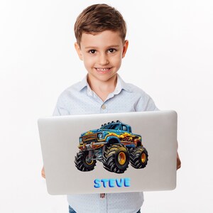 May include: A young boy is holding a laptop with a blue and yellow monster truck sticker on it. The sticker has flames on the side and the name "STEVE" is printed below the truck.