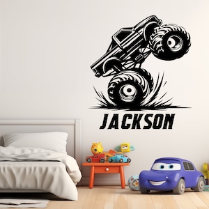 May include: Black and white silhouette of a monster truck with the name "JACKSON" printed below. The truck is on a white wall with a bed and a toy car in the background.