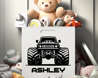 May include: White storage bin with a black monster truck graphic and the name "ASHLEY" in bold letters. The bin is filled with toys, including a teddy bear, a plush animal, and various toy cars. The bin has a handle for easy carrying.