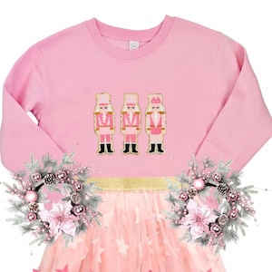 May include: Pink long-sleeved sweatshirt with three pink and gold nutcracker appliques. A pink tulle skirt with pink star appliques. Two pink and silver wreaths with pine branches and ornaments.