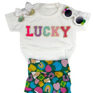 May include: A white short-sleeved t-shirt with the word "LUCKY" in rainbow glitter letters. The shirt is paired with green and white patterned shorts and a pair of sunglasses with the words "Lucky Charm" on the side.