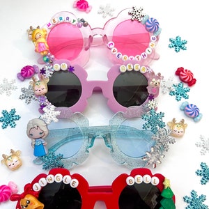 May include: Four pairs of colorful sunglasses with festive Christmas themes. The sunglasses have different colored frames and lenses, and each pair features a different Christmas-themed decoration, such as a reindeer, a snowflake, a bell, and a Christmas tree. The sunglasses are perfect for adding a festive touch to any outfit.