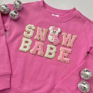 May include: Pink sweatshirt with the words "SNOW BABE" in white and gold letters with a pink and white snowman applique in the center.