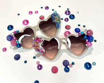 May include: A pair of white heart-shaped sunglasses with pink and blue glitter accents. The sunglasses are decorated with small pink, blue, and white beads.