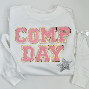 May include: White long-sleeved shirt with pink and gold lettering that says "COMP DAY" and a silver glitter star.