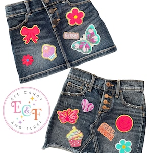 Könnte beinhalten: Zwei Jeansröcke mit bunten Patches. Der obere Rock hat Patches mit einer Blume, einer Schleife, einem Cupcake, einem Schmetterling und dem Wort "Boss". Der untere Rock hat Patches mit einem Herzen, einem Schmetterling, einem Cupcake, einem Kreis, einer Blume und den Worten "Girl Power".