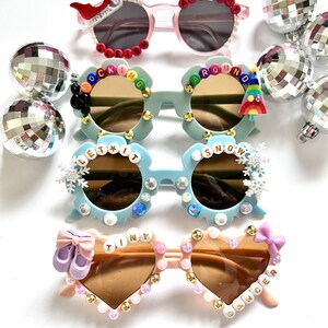 May include: Four pairs of decorative sunglasses with various embellishments. One pair is pink with the text "Sleigh All Day", another is light blue with the text "Rocking Around", a third is light blue with the text "Let It Snow", and the last is peach with the text "Tiny Dancer".