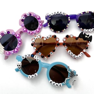 May include: Four pairs of Halloween themed sunglasses with different colored frames and embellishments. The sunglasses have phrases like "Boo", "Wicked", "Let's Go Ghouls", "Bad to the Bone", and "Lil' Cute" on them.