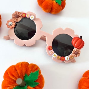 May include: Pink flower-shaped sunglasses with black lenses and a beaded word "HEY" on one side and "PUMPKIN" on the other. The sunglasses are surrounded by small pumpkins.