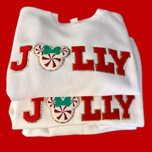May include: White sweatshirts with the word "JOLLY" in red letters with gold trim. The "O" is a Mickey Mouse head with a candy cane design and a green bow. The sweatshirts are folded on a red background.