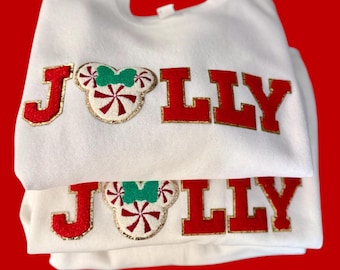 May include: White sweatshirts with the word "JOLLY" in red letters with gold trim. The "O" is a Mickey Mouse head with a candy cane design and a green bow. The sweatshirts are folded on a red background.