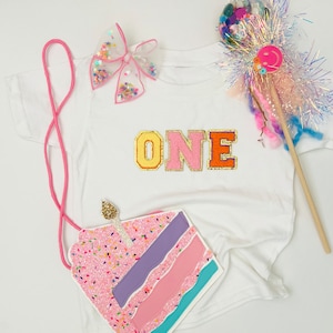 May include: A white t-shirt with the word "ONE" in colorful letters and a pink and blue glitter cake appliqué. The shirt is decorated with a pink bow and a sparkly wand.