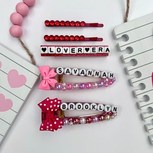 Puede incluir: Tres pinzas para el pelo de color rosa y rojo con los nombres "LOVERA", "SAVANNAH" y "BROOKLYN" escritos en letras blancas. Cada pinza tiene un lazo rosa y está decorada con cuentas rosas y blancas.