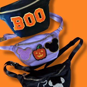 May include: Three Halloween-themed fanny packs in black, purple, and navy blue. The navy blue pack has the word "BOO" in orange letters. The purple pack has a purple Mickey Mouse head, a jack-o'-lantern, and a black Mickey Mouse head. The black pack has a witch hat and a Mickey Mouse head.