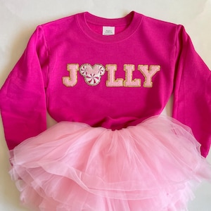 May include: A pink sweatshirt with a gold glitter "JOLLY" design featuring a candy cane Mickey Mouse head. The sweatshirt is paired with a pink tulle tutu.