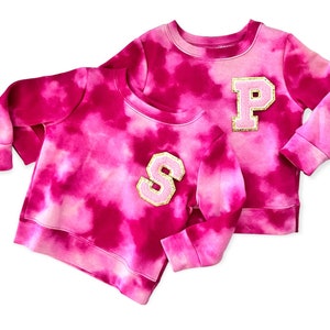May include: Two pink tie-dye sweatshirts with gold glitter letters. The sweatshirt on the left has a large "S" on the front and the sweatshirt on the right has a large "P" on the front.