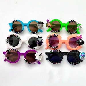 May include: Ten pairs of colorful plastic sunglasses with Halloween-themed decorations and phrases like "Lil Monster", "Magnificent Babe", "You Go Ghoul", "Sugar Skull", and "Cutie Pie".