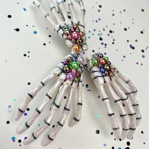 May include: Two white skeleton hand hair clips with iridescent rainbow colored beads. The clips are on a white background with iridescent glitter.