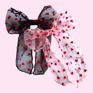 May include: Two hair bows with long tails. One bow is black with red hearts and the other is pink with red hearts.