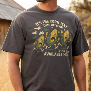 May include: Dark gray t-shirt with a humorous graphic. The design features three cartoon corn characters running away from disembodied hands, with the text "IT'S THE CORN-IEST TIME OF YEAR" and "FRESH CORN AVAILABLE NOW".