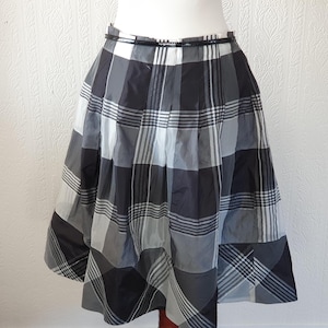 May include: A knee-length skirt with a black, white, and gray plaid pattern. The skirt has a pleated design and a black waistband. The fabric appears to be lightweight, suitable for warmer weather. The skirt is displayed on a mannequin.