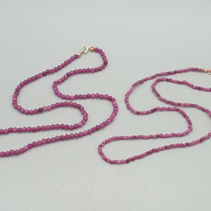 Può includere: Due delicate collane con piccole perline sfaccettate color magenta. Le collane hanno chiusure color oro. Le collane sono esposte su una superficie grigio chiaro. Gioielli.