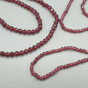 May include: Several strands of faceted, deep red gemstone beads. The beads are strung together to create necklaces or bracelets. The beads are small and have a sparkling, polished appearance. The background is a plain, light color.